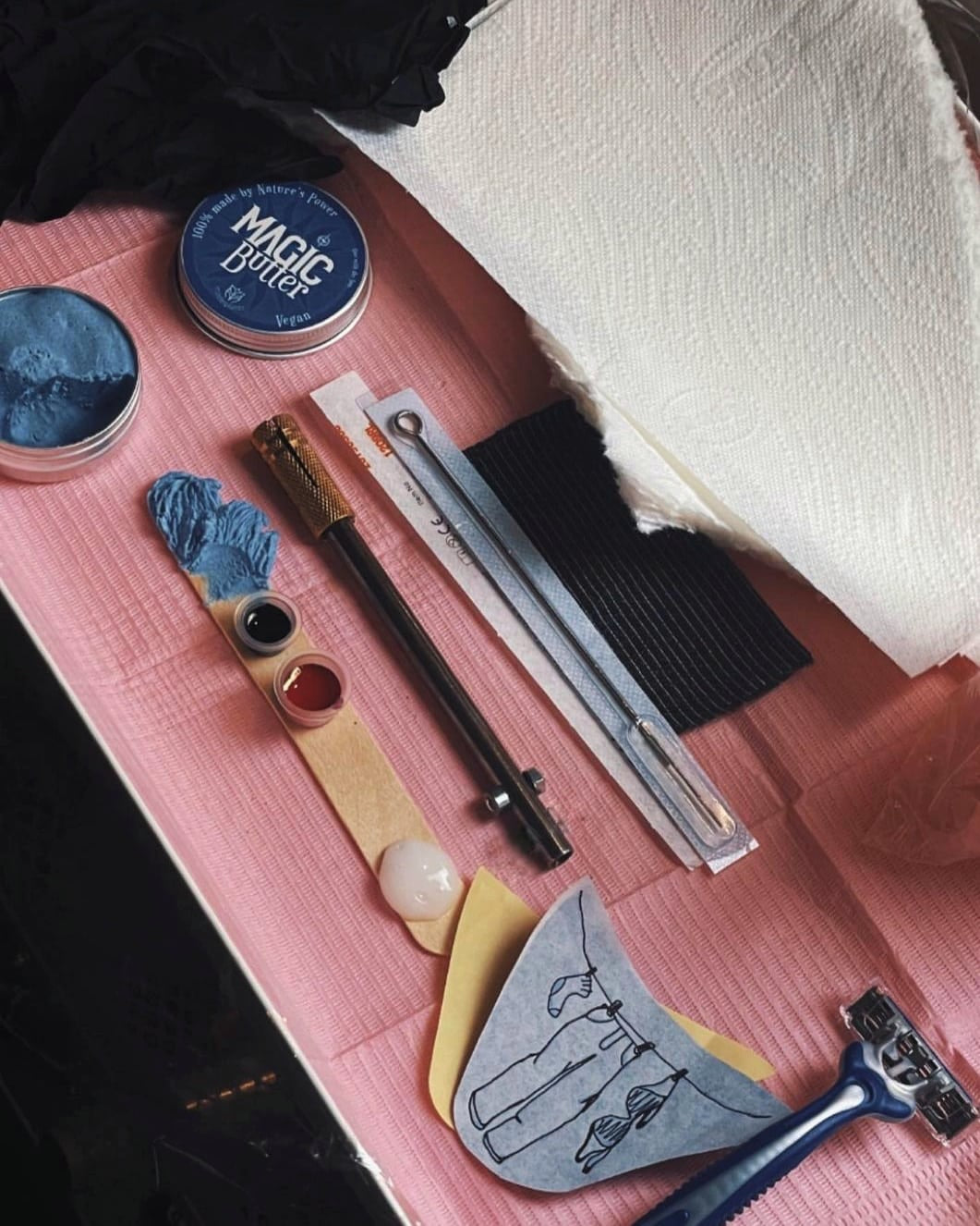 Collection of Handpoke tattooing tools and products on a pink surface, the Powk tool is in the middle and other tattoo things are around the side of it including, tattoo needles 